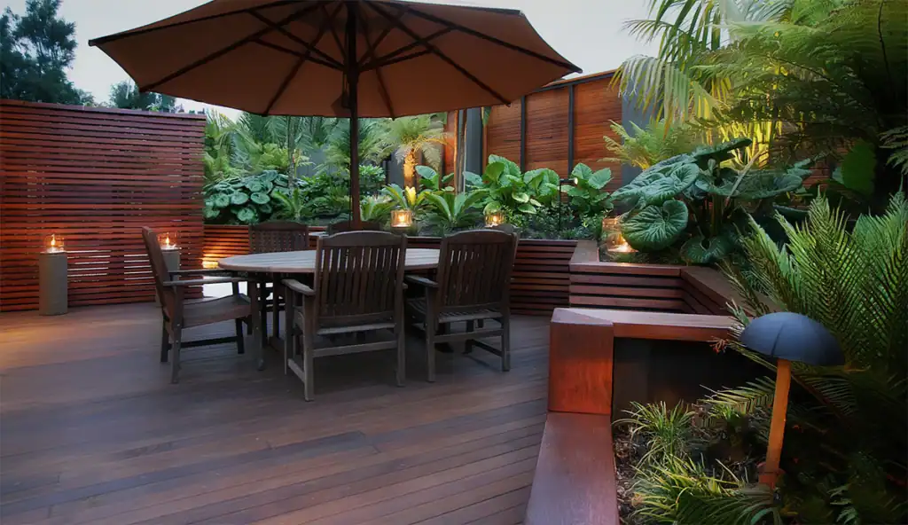 Modern garden design in Birkenhead featuring timber decking with built-in seating and timber slat screens surrounded by lush tropical shade loving plants.