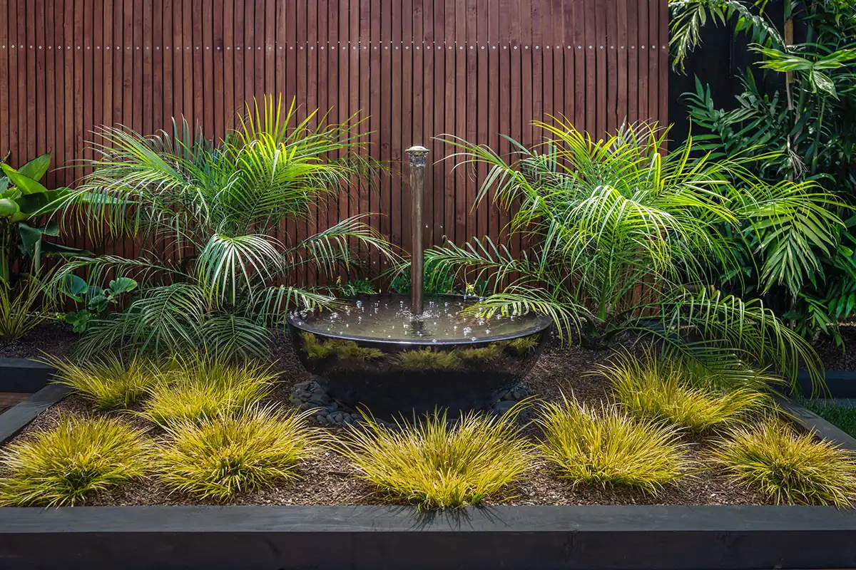 Display garden showcasing large bowl shaped water feature with copper spout, nestled in amongst plants with a vertical timber slat screen behind it.